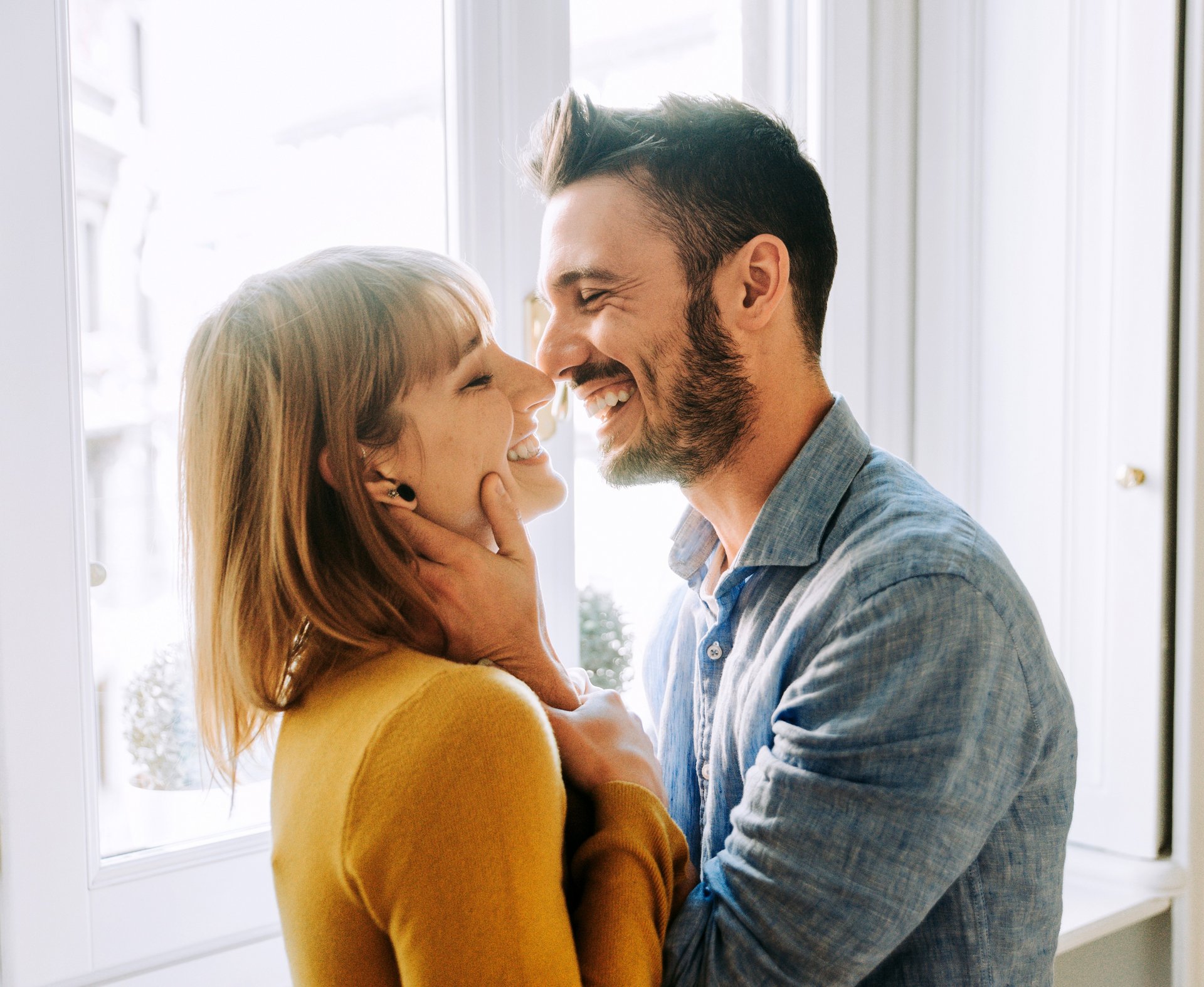 Ein Paar, das sich anlächelt, steht vor einem hellen Fenster. Die Frau trägt einen gelben Pullover, der Mann ein blaues Hemd.